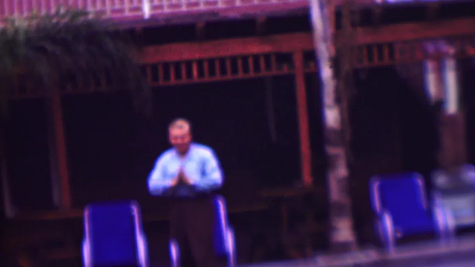 The adult man faces the camera with his hands together in a brief polite gesture, standing near blue deck chairs and the shaded porch area.