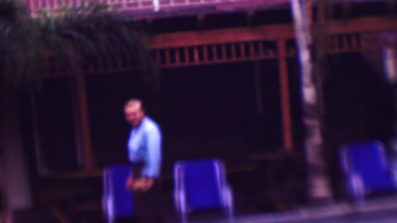 In this 1961 San Diego home-movie frame, an adult man walks beside a pool in front of blue lounge chairs, palm trunks, and a dark wood-railed exterior.