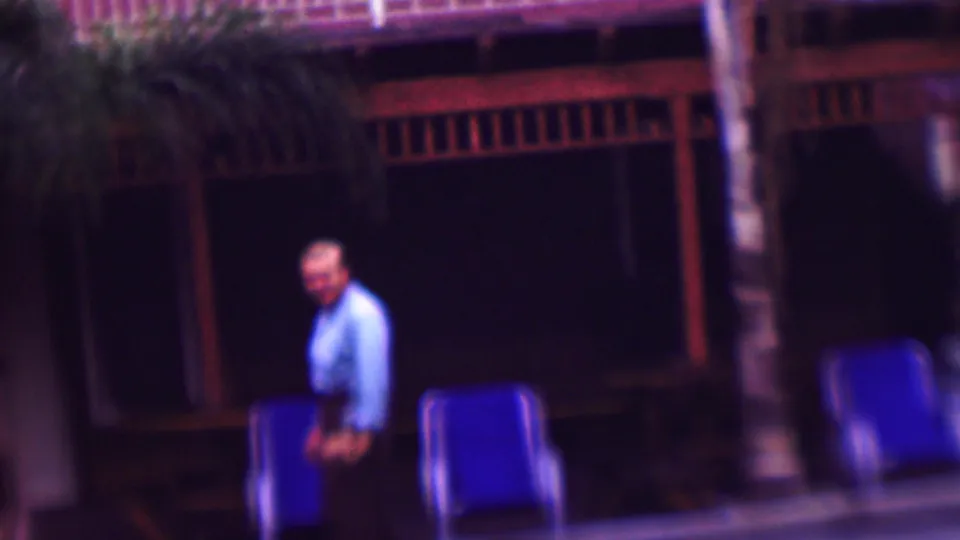 In this 1961 San Diego home-movie frame, an adult man walks beside a pool in front of blue lounge chairs, palm trunks, and a dark wood-railed exterior.