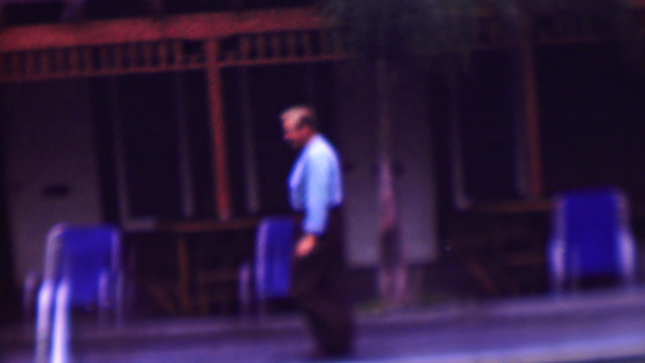 The man continues along the poolside area, passing blue lounge chairs in front of a shadowed building with wooden railings and palm trunks.