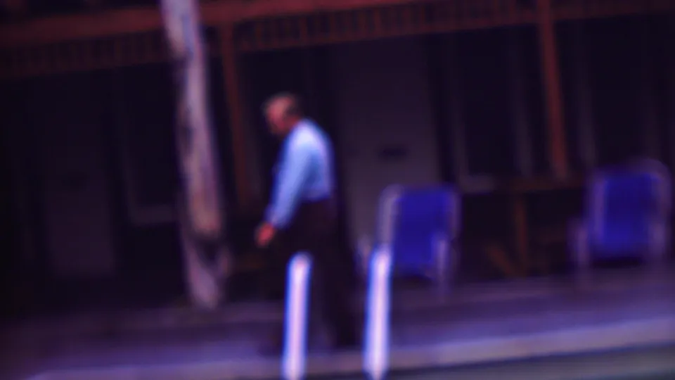 A soft-focused frame shows the adult man moving along the pool edge, with blue lounge chairs, a palm trunk, and a dark exterior wall behind him.
