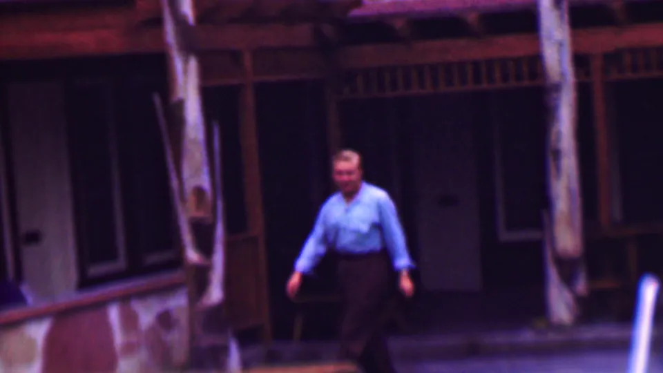 The man faces more toward the home-movie camera near the pool, framed by blue lounge chairs, palm trunks, and a shadowed wood-railed exterior.
