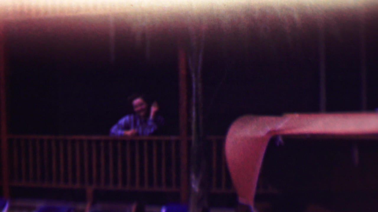 In this 1961 San Diego home-movie frame, a man in a striped shirt appears on a wooden porch at night, smiling toward the camera with one hand raised.