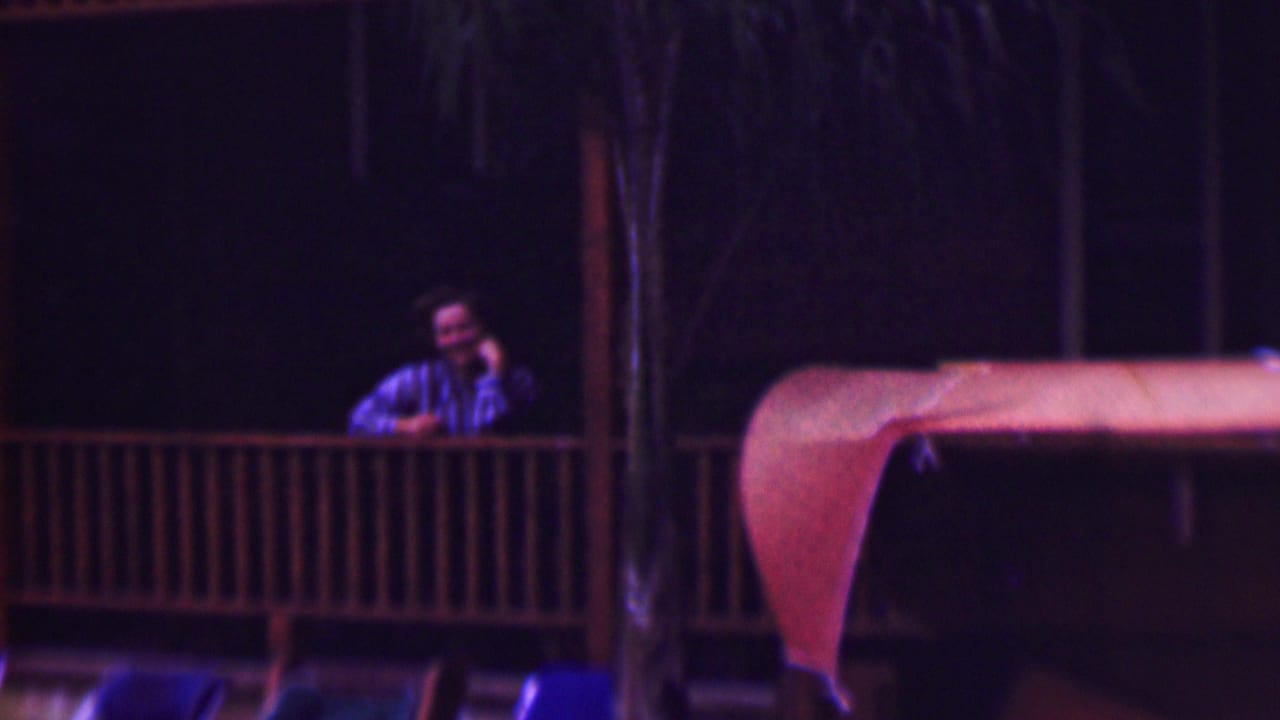 Man on a porch smiles toward the camera beside a railing and dark trees.