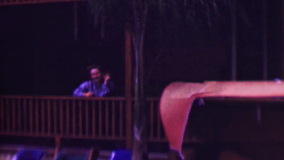 The frame shows the same man in a striped shirt waving from a dim wooden porch, with strong 8mm color shift and a dark outdoor background.