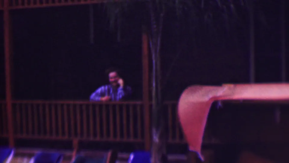 Near the end of the clip, the man is framed behind the porch railing at night, with dark trees, vertical posts, and a red-orange roof or awning edge visible in the 8mm scan.