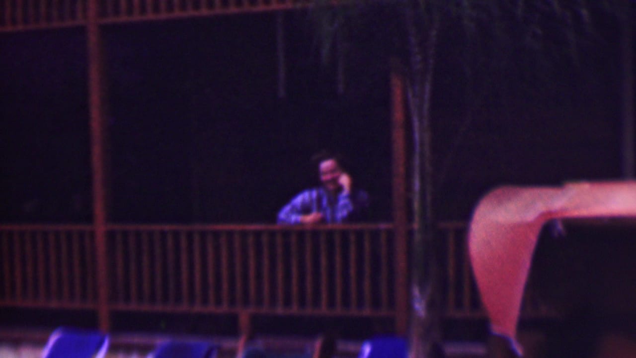A man in a striped shirt appears on an upper wooden balcony above a dim hotel pool area in supplied 1961 San Diego home-movie footage.