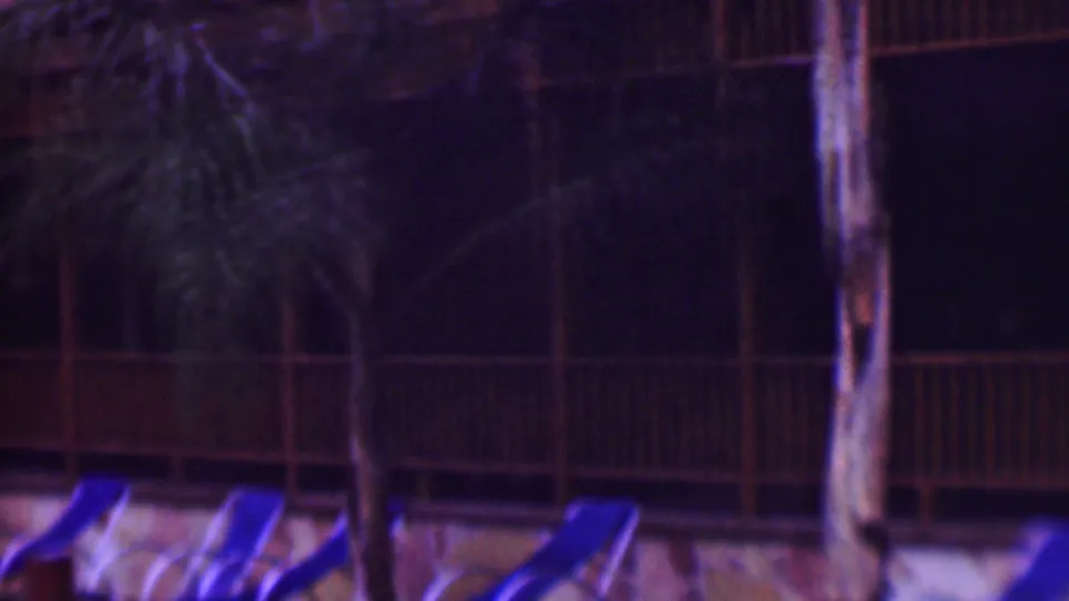 Palm trunks, wooden balcony railings, and blue lounge chairs frame a dark hotel pool area in this nighttime 8mm home-movie frame.