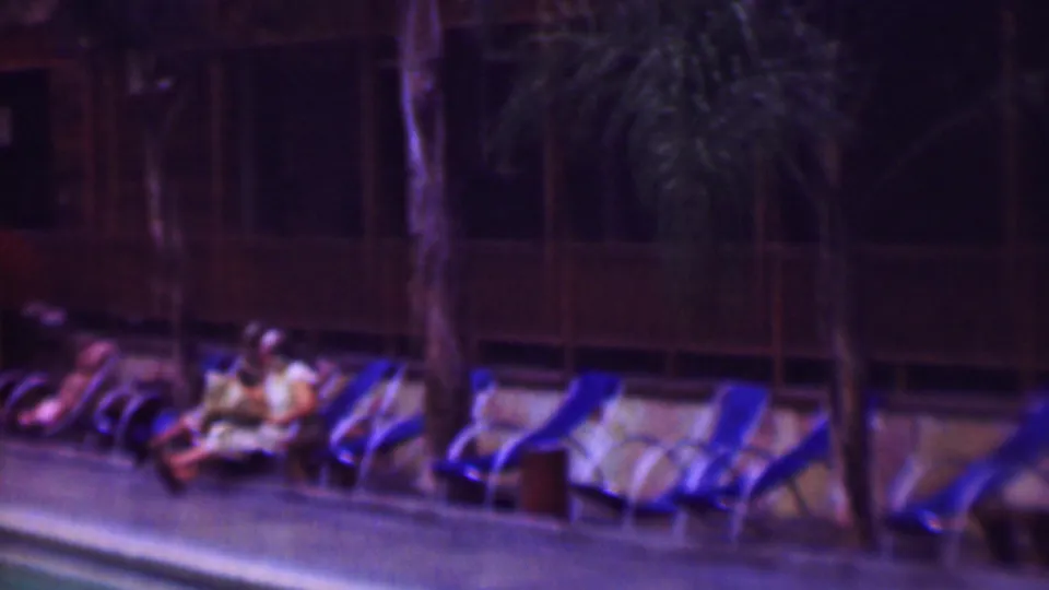Several people rest near the pool beside blue lounge chairs, with wooden hotel railings and palm trunks visible in the nighttime 1961 San Diego footage.
