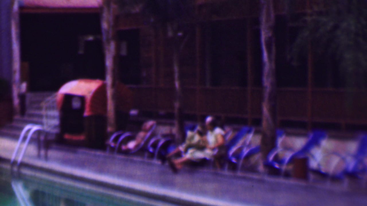 1961 home-movie frame from San Diego showing people relaxing on lounge chairs beside a swimming pool, with a ladder, palm trunks, and curtained building windows in the background.