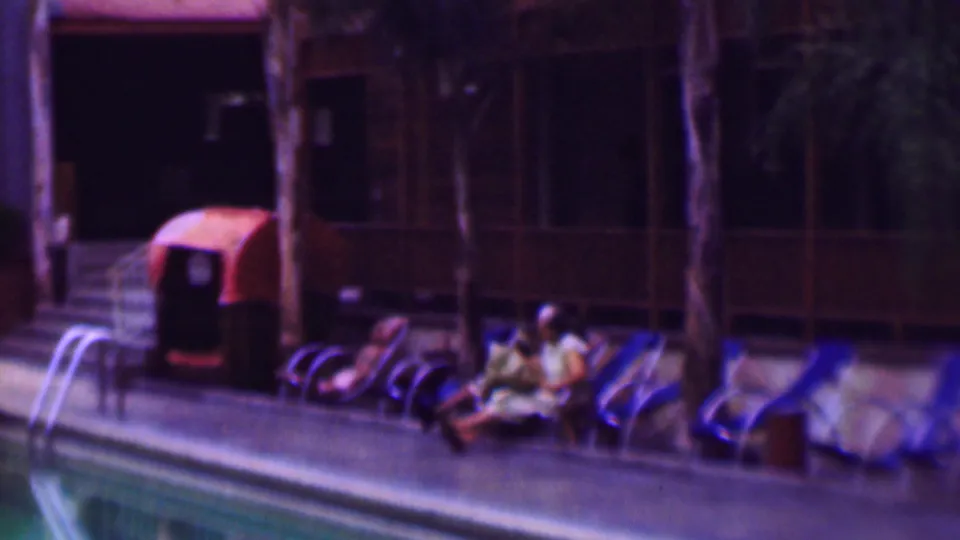 1961 home-movie frame from San Diego showing people relaxing on lounge chairs beside a swimming pool, with a ladder, palm trunks, and curtained building windows in the background.
