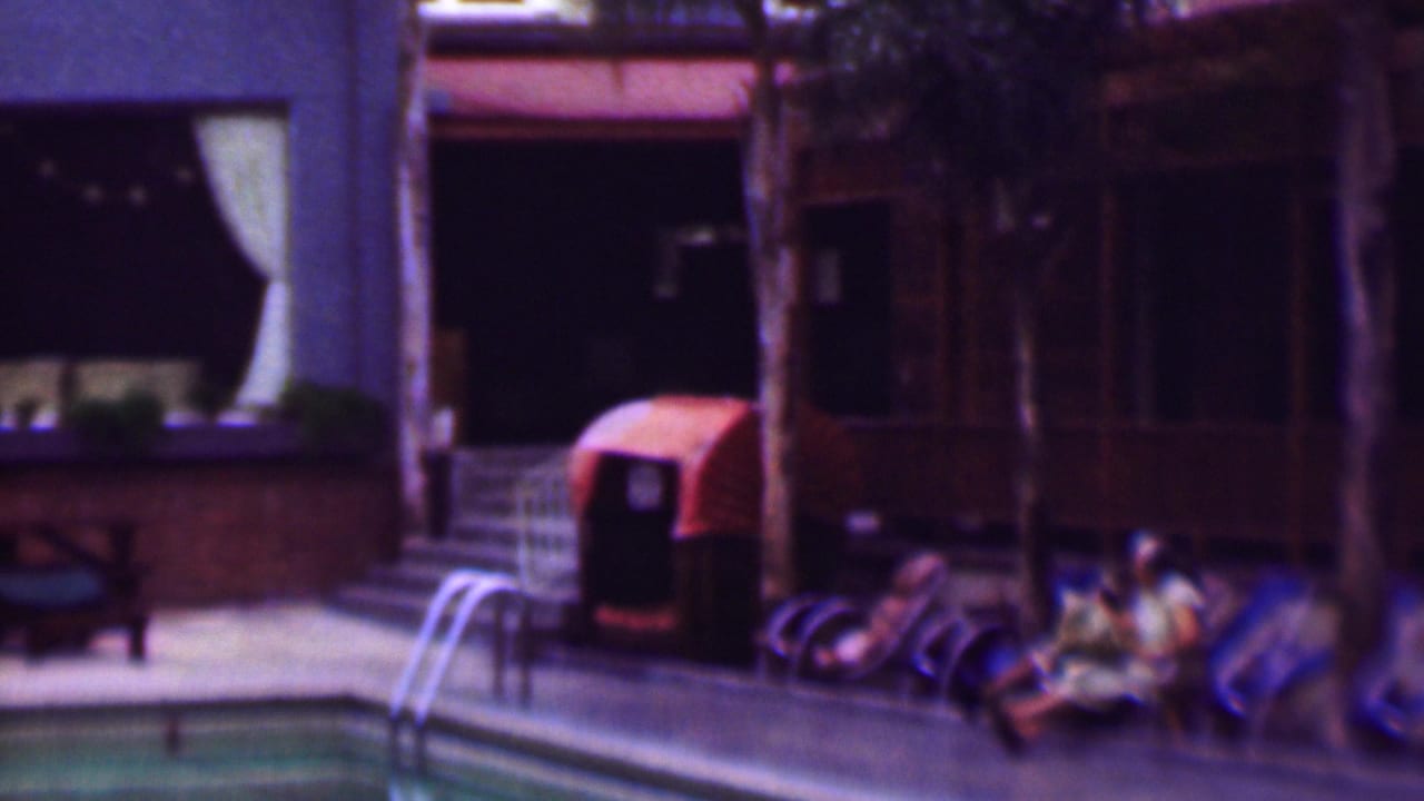 Lounge chairs and seated people beside a pool, with a ladder and curtained building windows nearby.