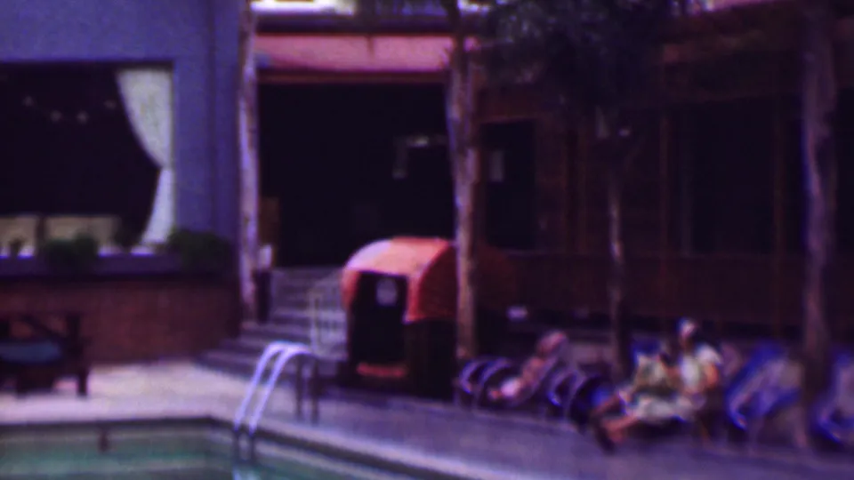 1961 San Diego poolside frame showing seated people near lounge chairs, a pool ladder, palm trunks, and a building with dark windows and curtains.
