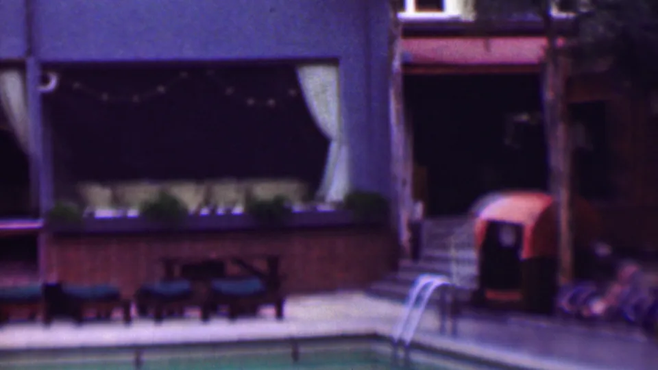 1961 San Diego home-movie frame showing a poolside patio with lounge chairs, small tables, a pool ladder, palm trunks, and curtained building windows.