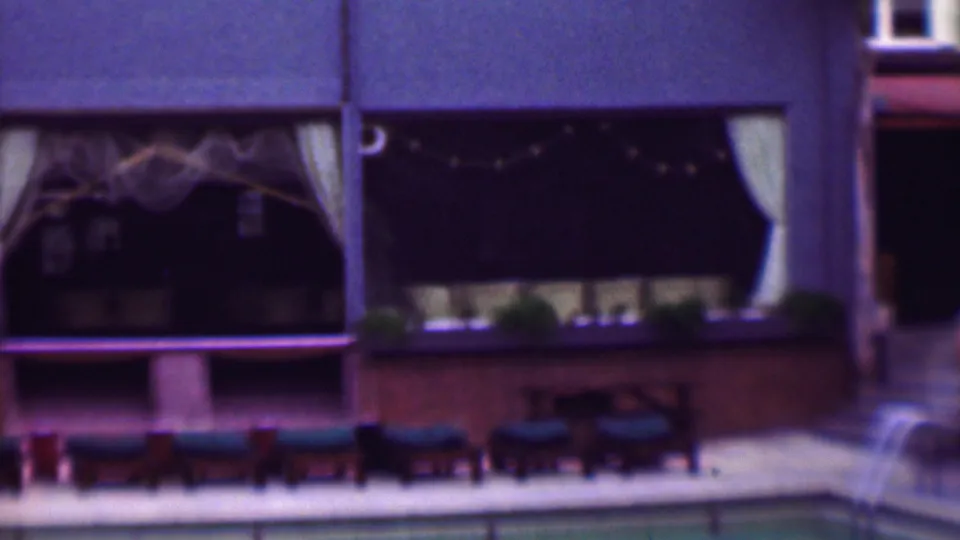 1961 San Diego poolside frame showing lounge chairs and tables along a patio, with palm trunks, string lights, and curtained windows across the building facade.