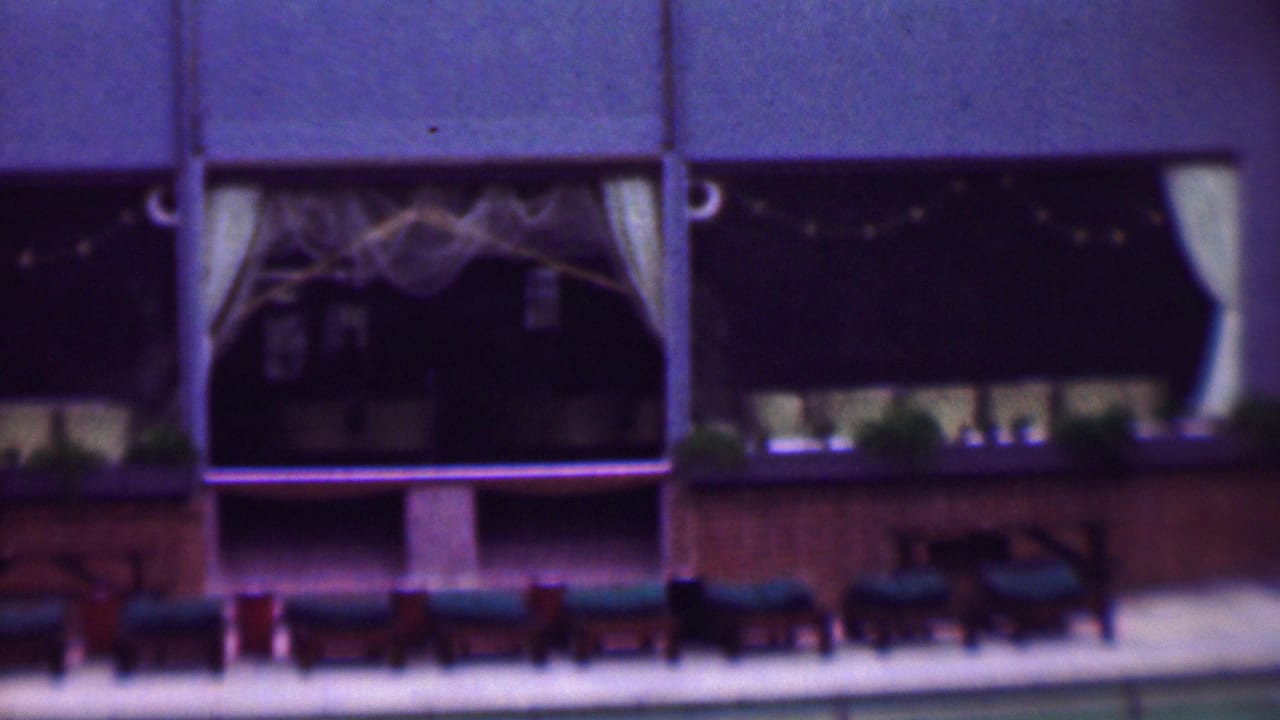 1961 San Diego home-movie frame showing a restaurant or hospitality exterior with dark window openings, pale curtains, string lights, and rows of poolside lounge chairs.