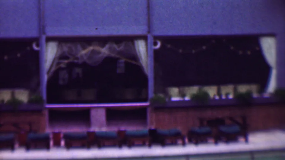 1961 San Diego home-movie frame showing a restaurant or hospitality exterior with dark window openings, pale curtains, string lights, and rows of poolside lounge chairs.