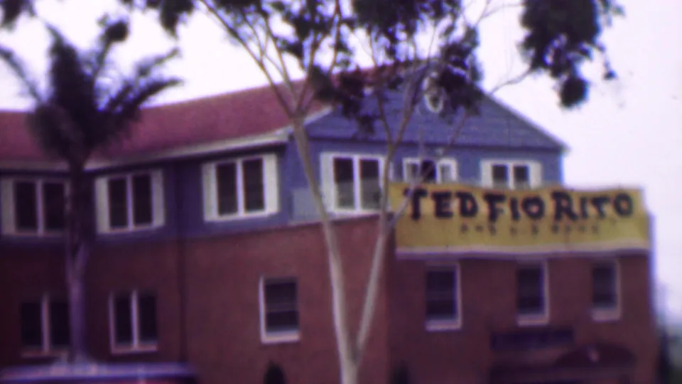 1961 San Diego home-movie frame showing a red-brown building exterior with palm trees, upper windows, and a yellow sign with partially legible restaurant lettering.