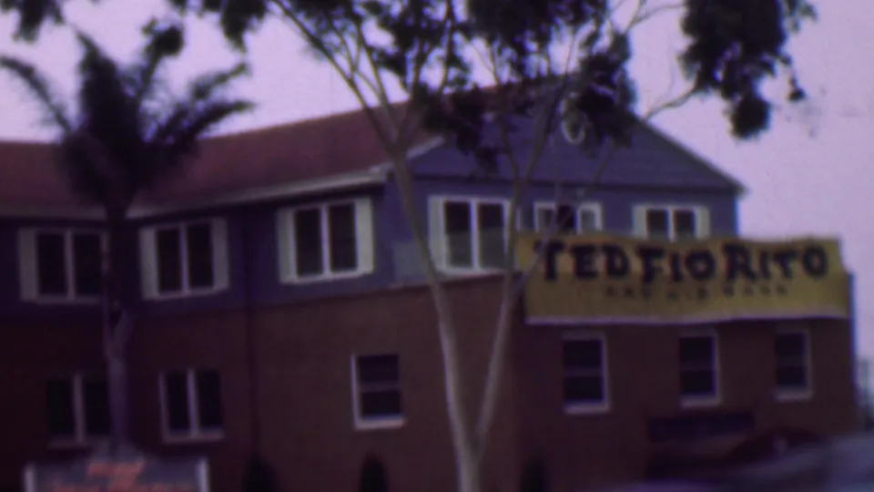 1961 San Diego exterior frame showing palm trees in front of a red-brown restaurant or hospitality building, with a yellow sign and multiple windows visible.