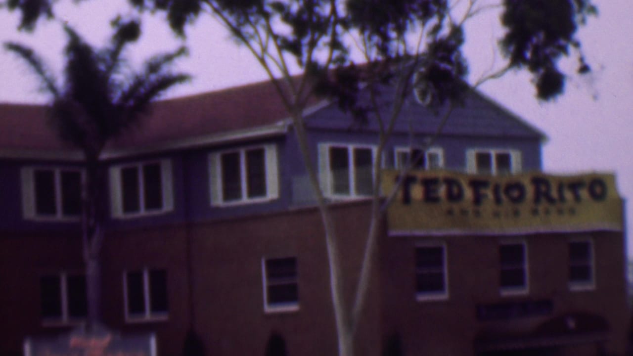 1961 San Diego home-movie frame showing a soft-focused two-level building with blue upper walls, brown lower walls, white windows, foreground branches, and a yellow Ted Fio Rito and His Band banner.