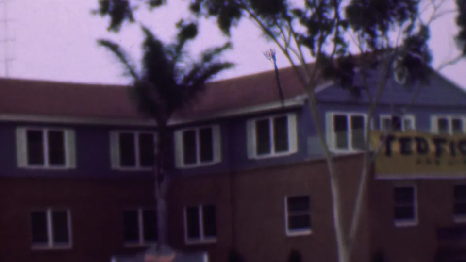 The frame shows the same 1961 San Diego exterior from a slightly shifted angle, with white-trimmed windows, foreground trees, and the yellow Ted Fio Rito banner cut off at the right edge.