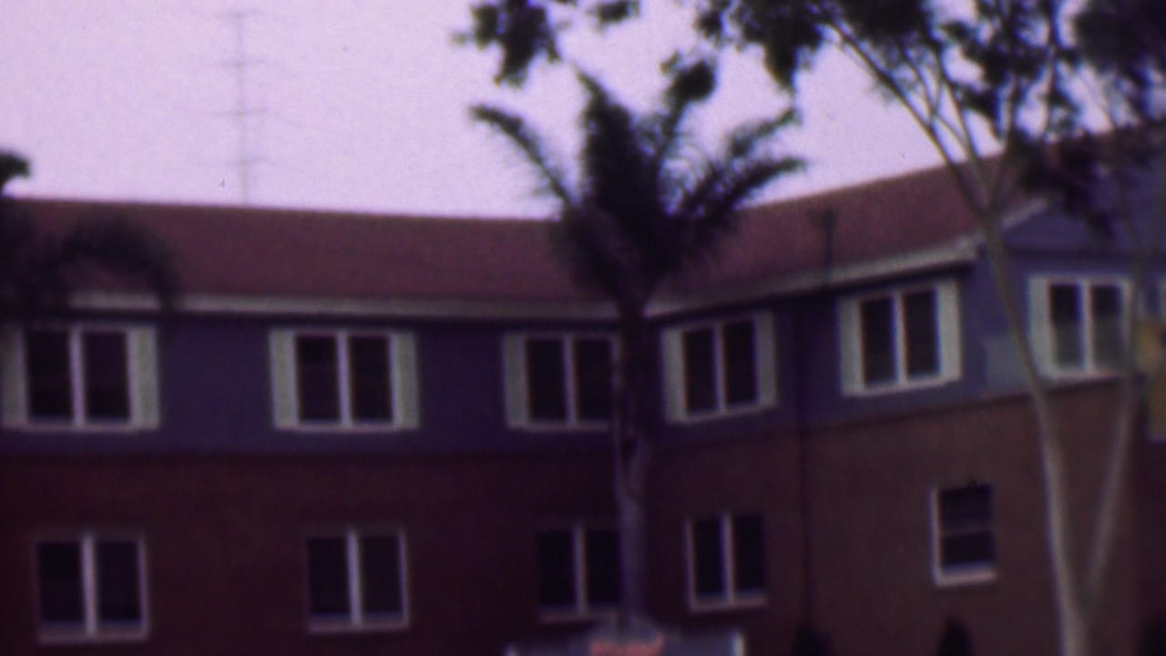 1961 San Diego home-movie frame showing a large red lower and blue upper building with white-framed windows, palm trees, and leafy branches crossing the view.