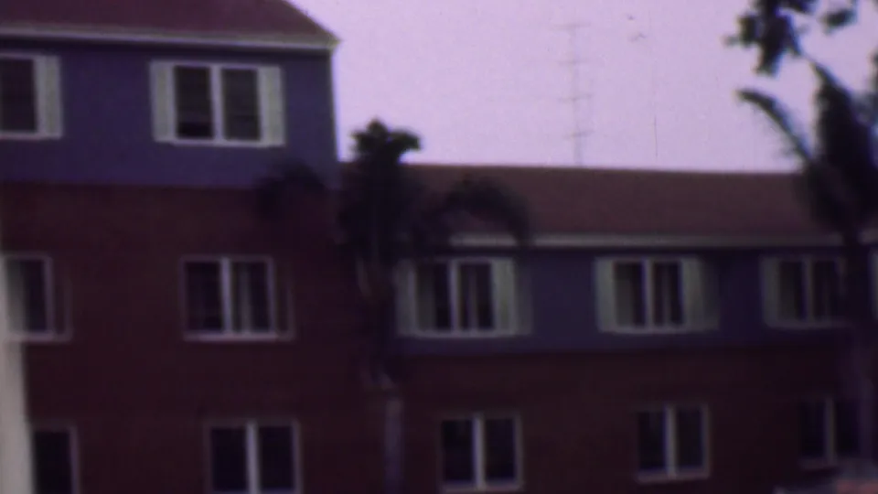 1961 San Diego home-movie frame showing the building from a slightly shifted angle, with repeated white-framed windows, palm trunks, dark foliage, and the red roofline.
