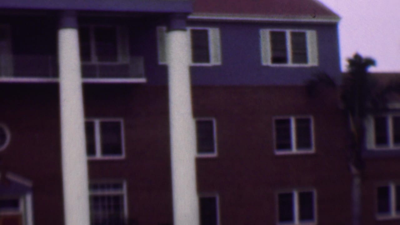 1961 San Diego home-movie frame showing the upper facade of a red brick building with tall white columns, windows, balcony areas, and a palm trunk near the center.
