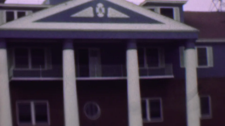 1961 San Diego frame showing a columned entrance section with a triangular gable, balcony railings, red brick walls, rectangular windows, and a small round window.