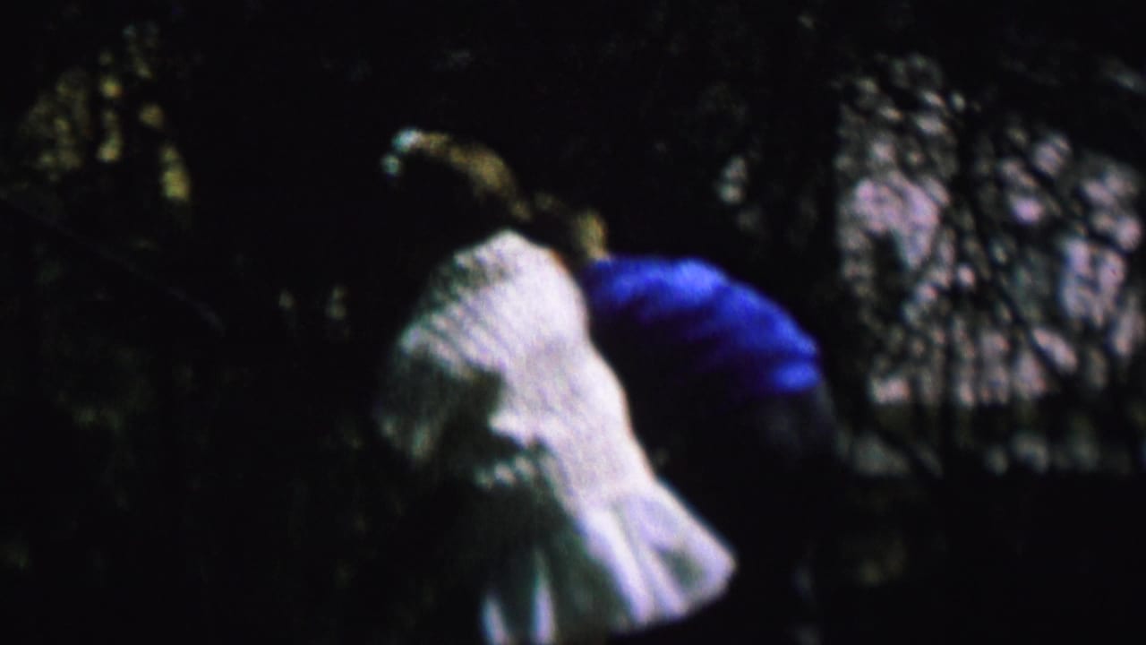 In this 1964 home-movie frame, a child in light clothing appears near shrubs or shadowed yard growth, with a blue garment visible beside them in the outdoor setting.