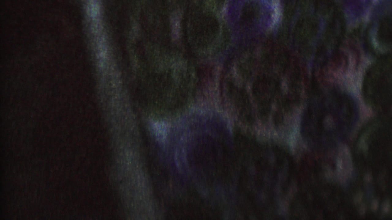 Blurred adult figure in dark clothing near another figure in a low-light scene.