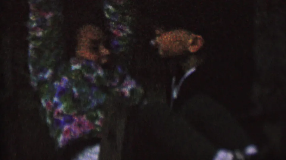 1962 8mm St. Paul frame showing a woman in a floral dress with her hands held or bound behind her back while a man in dark formal clothing stands close by.