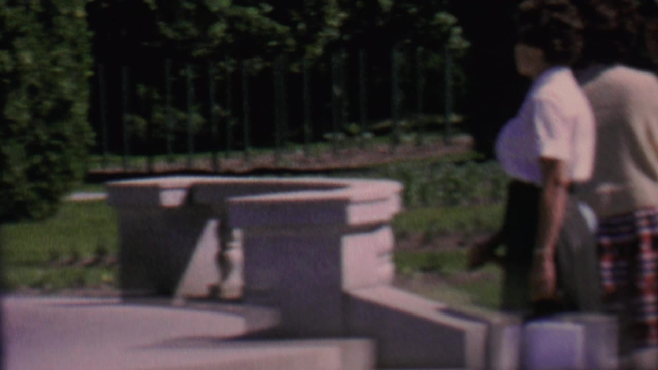 1962 home-movie frame from the supplied St. Paul context showing adults walking and sitting near a stone fountain in a landscaped park.