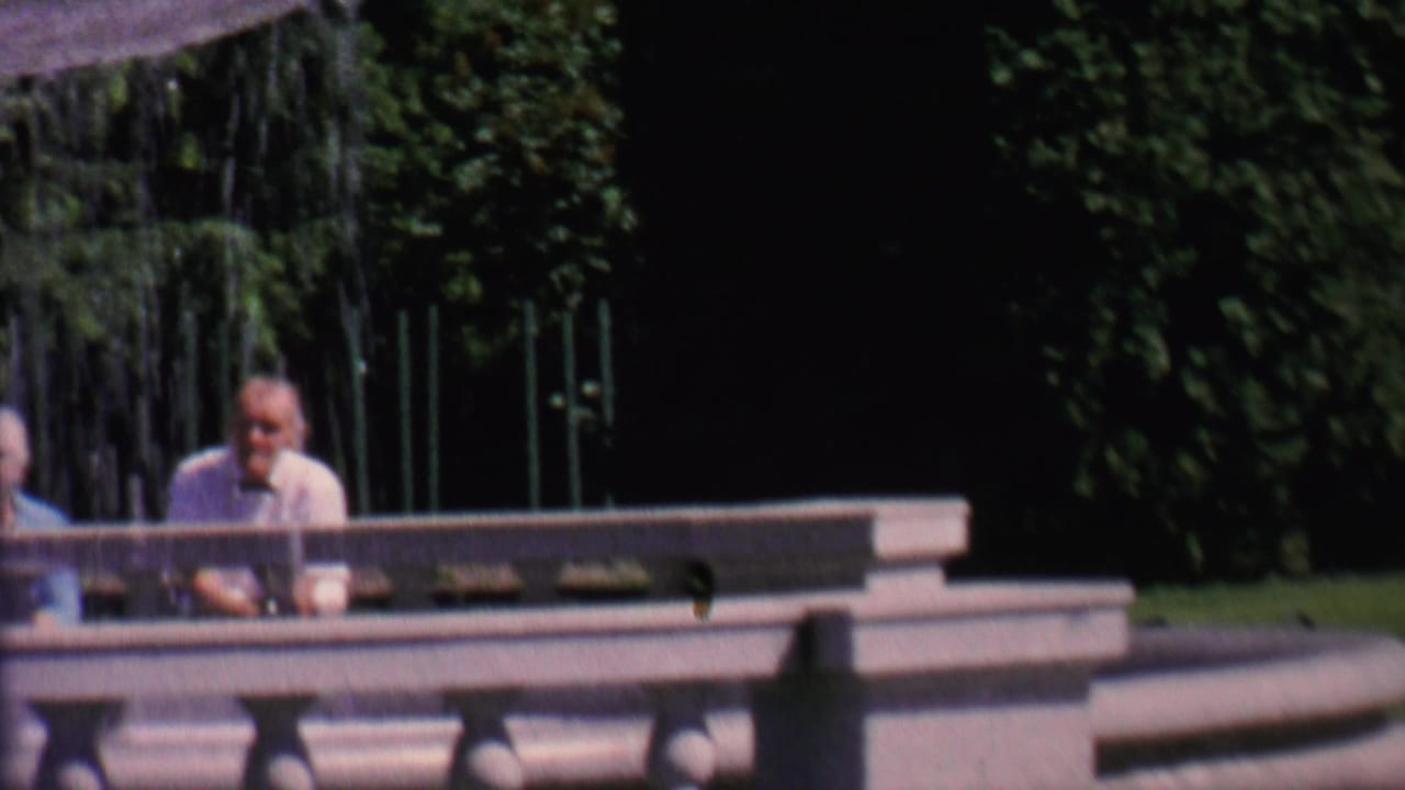Two adults in mid-century clothing walk in front of a fountain while a man sits behind a railing.