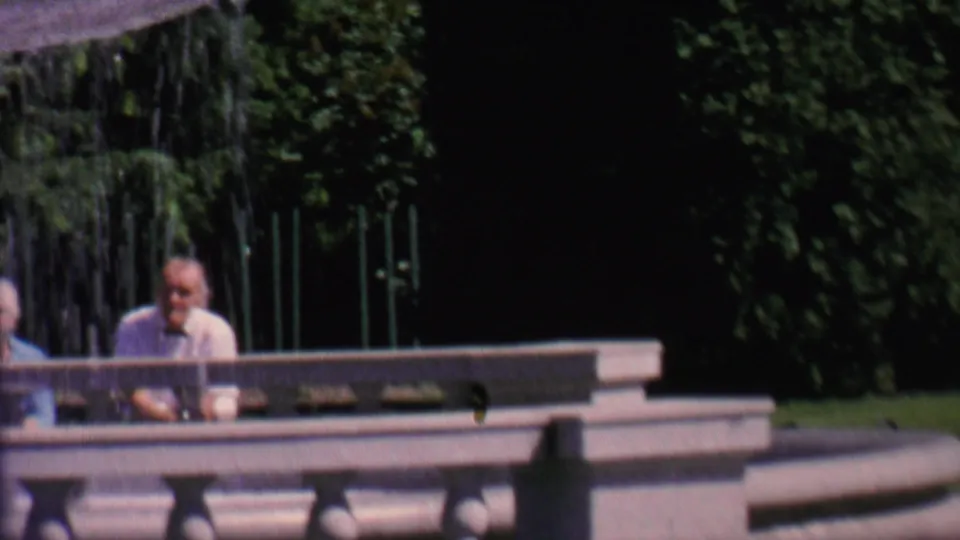 Two adults pass through the foreground near cascading fountain water as a seated man remains visible behind the low stone railing.
