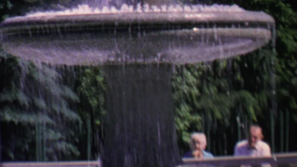A close home-movie view of a broad fountain bowl with water spilling down, with two seated adults partly visible beyond the fountain edge.