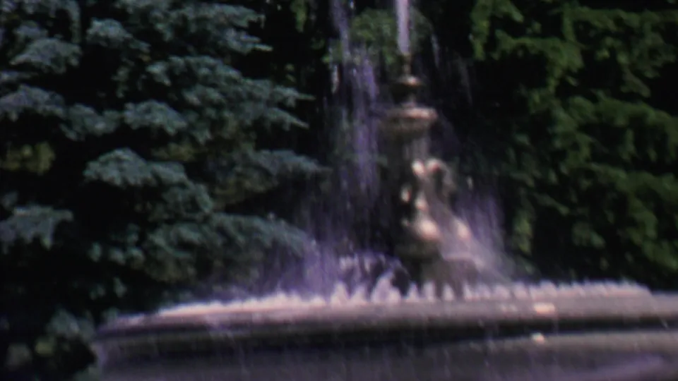 The tiered stone fountain fills the foreground with splashing water while two adults sit behind it against dense park greenery.