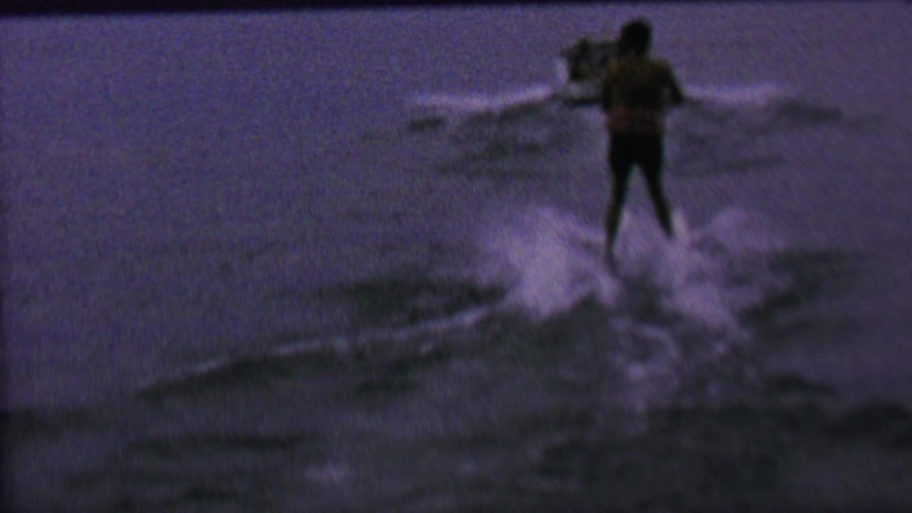 A distant water skier in dark trunks rides behind a speedboat on open water in metadata-supported 1962 United States home-movie footage.