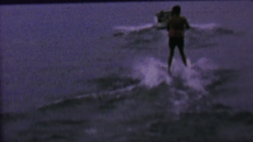 A distant water skier in dark trunks rides behind a speedboat on open water in metadata-supported 1962 United States home-movie footage.