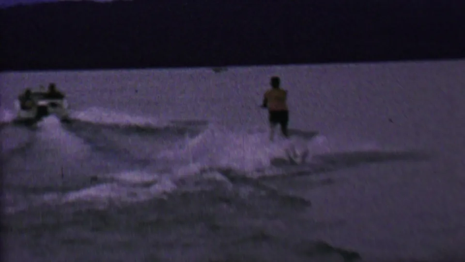 The skier continues behind the boat as white spray spreads across the lake surface in a distant 8mm home-movie view.
