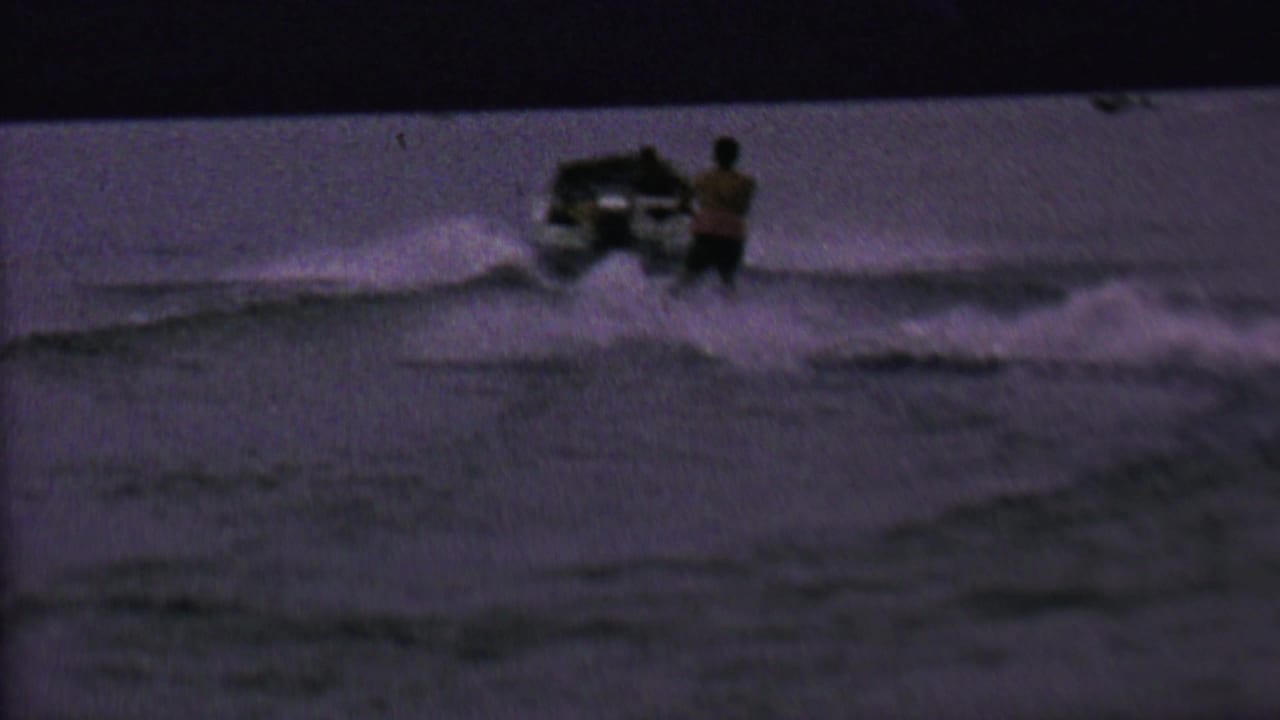 1962 United States 8mm frame showing a small speedboat and a beginner water skier near the wake in low evening light.