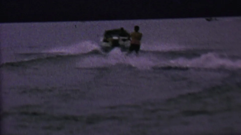 1962 United States 8mm frame showing a small speedboat and a beginner water skier near the wake in low evening light.