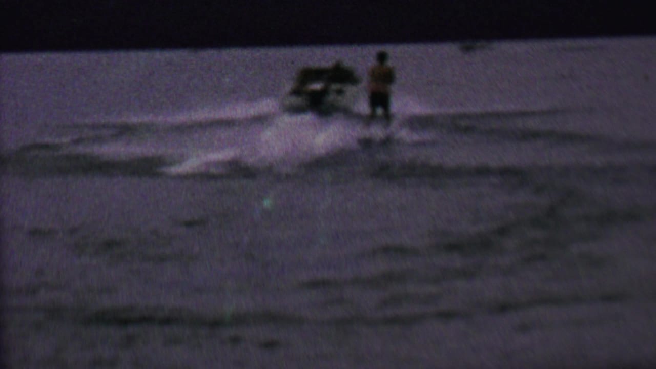 1962 home-movie frame from the United States showing a speedboat pulling a skier across a dim lake surface.