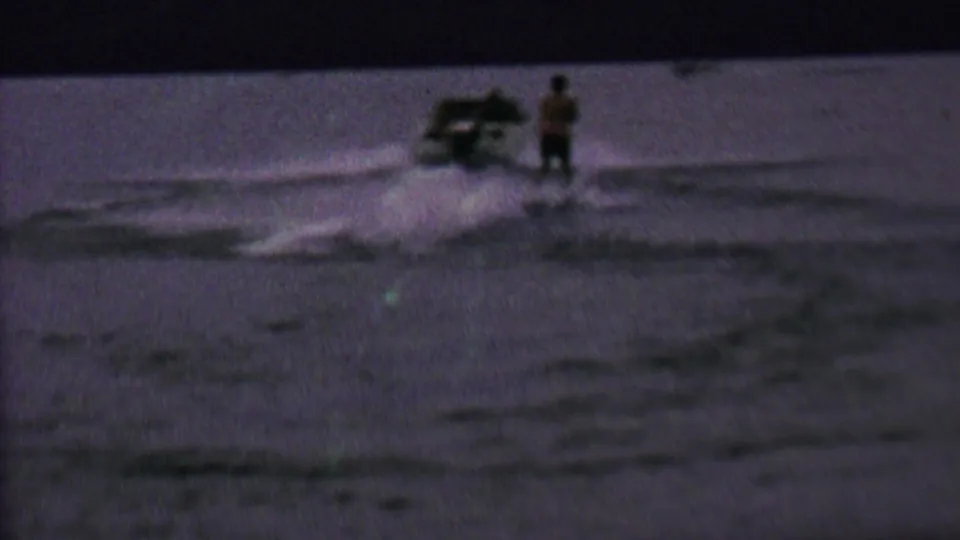 1962 home-movie frame from the United States showing a speedboat pulling a skier across a dim lake surface.