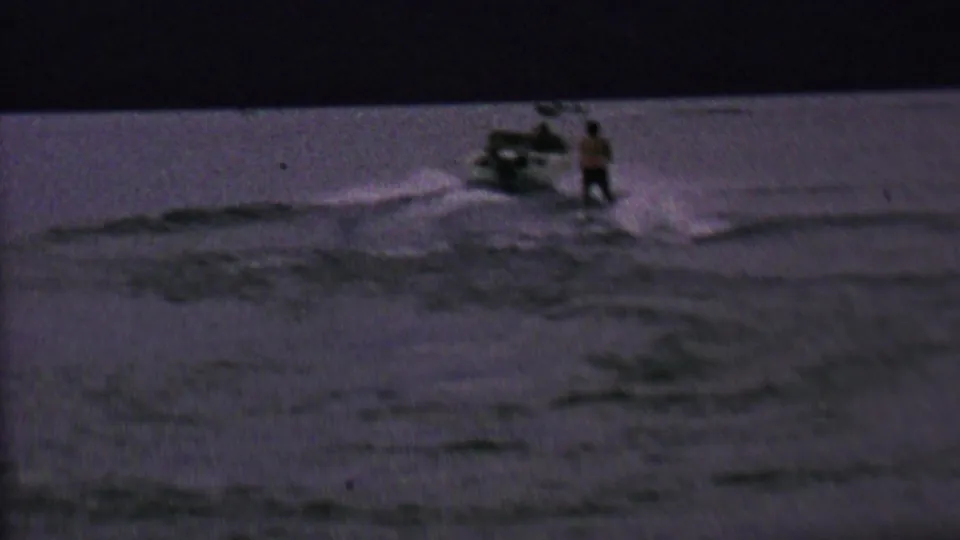 Grainy 8mm frame showing the skier near the wake as the small boat continues across the lake in low light.