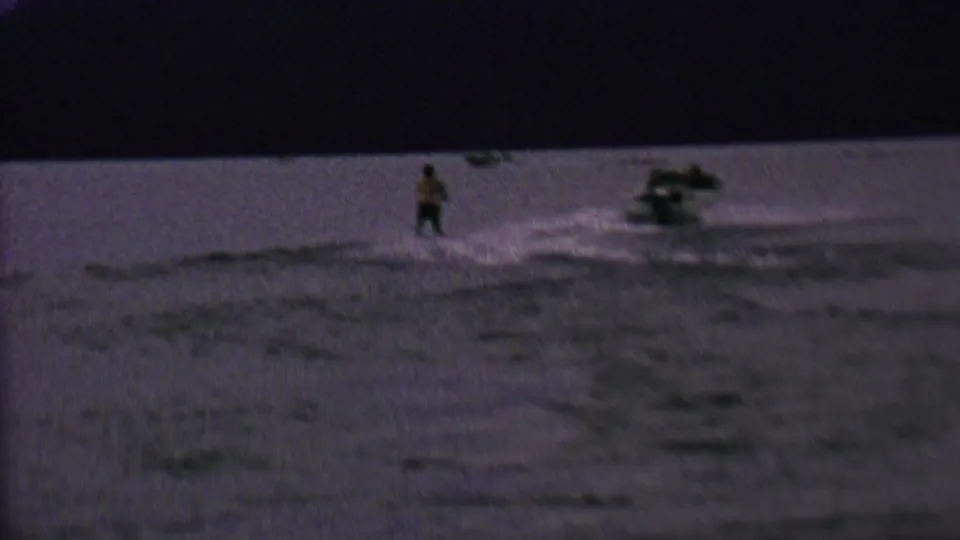 Wide 1962 home-movie frame showing a distant skier and speedboat with a foamy wake on dark lake water.