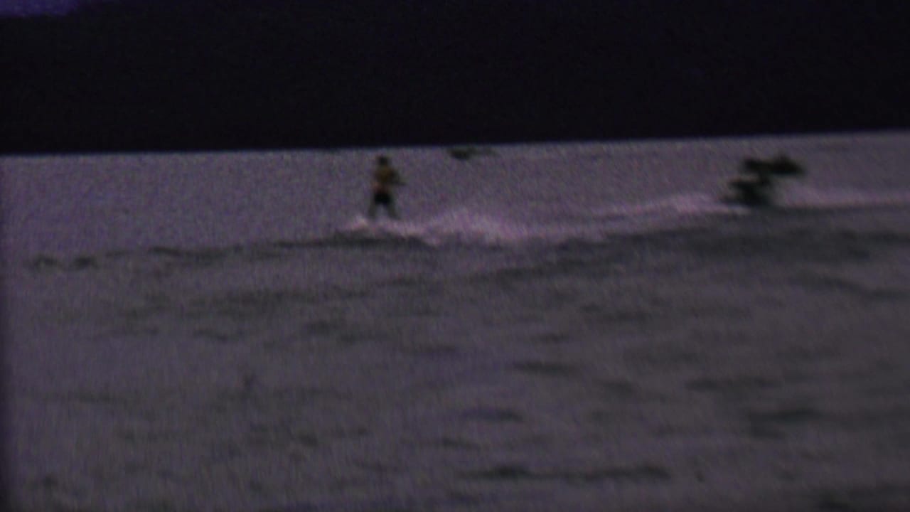 In 1962 home-movie footage associated with St. Paul, Minnesota, a distant male water skier moves across a dim lake behind a boat wake.