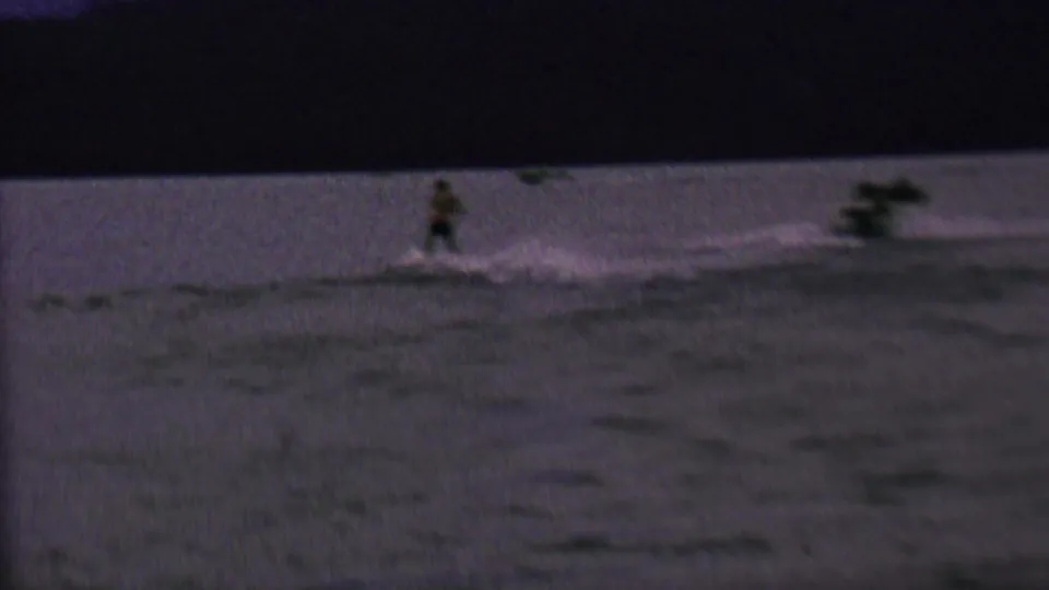 In 1962 home-movie footage associated with St. Paul, Minnesota, a distant male water skier moves across a dim lake behind a boat wake.