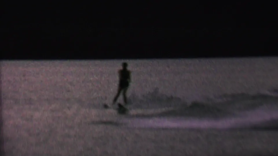 A silhouetted male water skier is pulled across the rippled lake surface, with the tow line and wake visible in low evening light.