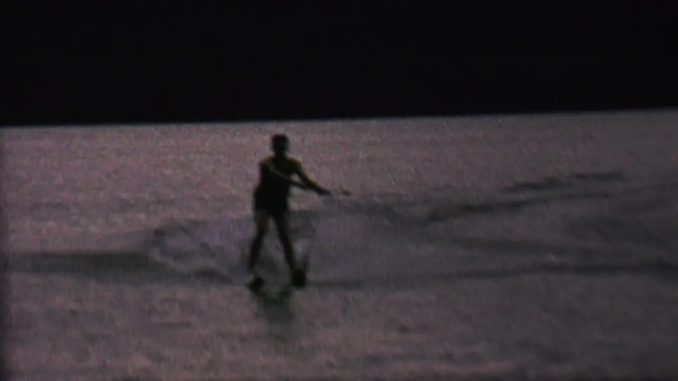 A silhouetted male water skier leans while holding the tow handle, with spray and wake spreading across the low-light lake scene.